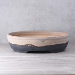 May include: A shallow, round ceramic bonsai pot featuring a two-tone design. The upper portion has a speckled, light beige finish, while the lower section is a solid, matte dark charcoal grey. The pot has a wide, open top and a flat base.