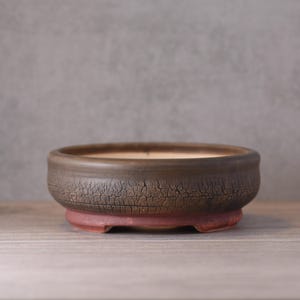May include: A brown ceramic bonsai pot with a cracked texture on the upper section and a red base. The pot is round and shallow, with a wide opening. The pot is on a wooden surface.