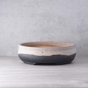 May include: A shallow, round ceramic bonsai pot featuring a two-tone glaze. The lower portion is a matte charcoal black, while the upper section is a speckled, light beige cream colour with a distinct, uneven horizontal transition line.