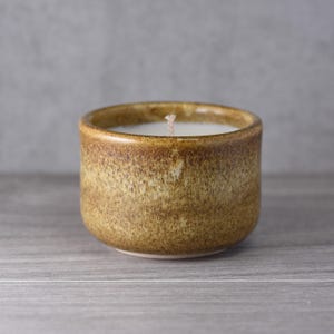 May include: A small, round candle in a ceramic container. The candle has white wax and a single wick. The container is a warm, earthy brown with a textured surface, giving it a rustic appearance. The candle sits on a wooden surface.