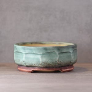 Peut inclure: Un pot à bonsaï rond en céramique turquoise avec un motif géométrique texturé. Le pot repose sur une base brun rougeâtre avec un intérieur jaune. Le pot est conçu pour planter de petits arbres ou plantes.