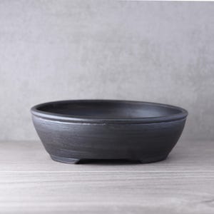 Op de afbeelding: Een ondiepe, ovale bonsaipot met een matte zwarte afwerking. De container heeft een glad, licht getextureerd oppervlak, een brede opening en een stevige basis met kleine voetjes, ontworpen voor het planten van kleine bomen of struiken.
