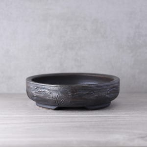 May include: A round, dark brown ceramic bonsai pot with a low profile and a textured, weathered exterior surface. The vessel features a wide, open top and is supported by small, integrated feet at the base, showcasing a rustic, earthy finish.
