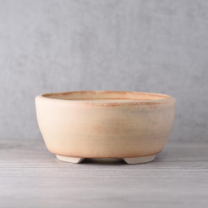 Può includere: Una piccola ciotola in ceramica beige con forma arrotondata e tre piedini. La ciotola ha una consistenza leggermente ruvida e un bordo più scuro. Il colore neutro e il design semplice suggeriscono un utilizzo per piante o come oggetto decorativo.