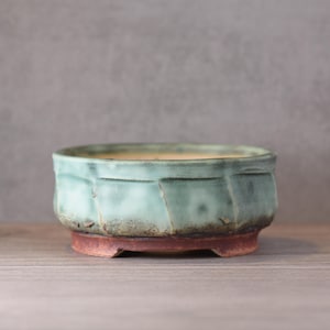 Może przedstawiać: Ręcznie wykonany doniczka do bonsai o owalnym kształcie. Doniczka ma glazurę w kolorze zieleni morskiej na teksturowanej powierzchni i czerwonobrązową podstawę. Jest podniesiona na małych nóżkach i nadaje się do małych roślin.