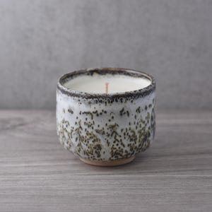 Könnte beinhalten: Eine Kerze in einem gesprenkelten weiß-braunen Keramikbecher. Die Kerze hat weißes Wachs und einen einzelnen Docht. Der Becher hat eine runde Form und steht auf einem kleinen, unglasierten Sockel. Der Rand des Bechers ist dunkelbraun.
