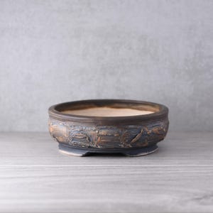 May include: A round, low-profile ceramic bonsai pot featuring a dark, textured exterior with weathered brown and charcoal grey tones. The interior of the vessel is a smooth, light beige colour, and the base is supported by small, integrated feet.