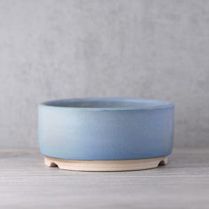 May include: A round, pale blue ceramic bowl with a cream base. The bowl has a smooth, matte finish and rests on three small feet. The interior of the bowl is a slightly darker shade of blue.