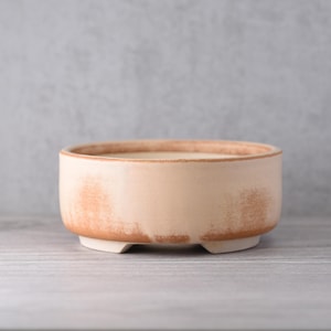 Op de afbeelding: Een kleine, ronde, beige keramische plantenbak met een verweerde afwerking. De plantenbak heeft een laag profiel met een brede opening en staat op drie kleine voetjes. De rand en de voetjes hebben een donkerdere, roodbruine kleur.
