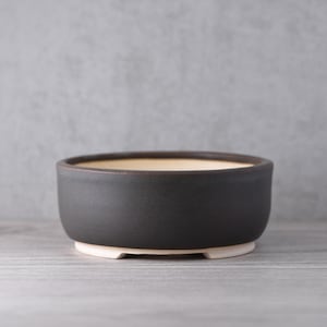 Könnte beinhalten: Ein runder, schwarzer Bonsai-Topf aus Keramik mit hellbeigem Innenraum und weißem Sockel. Der Topf hat ein schlichtes, modernes Design, perfekt für kleine Pflanzen. Der Topf hat einen Durchmesser von ca. 15 cm und eine Höhe von 7,6 cm.