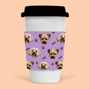 Könnte beinhalten: Ein weißer To-Go-Kaffeebecher mit einer lilafarbenen Hülle, die ein sich wiederholendes Muster aus Hundegesichtern, Pfotenabdrücken und Knochenformen zeigt.