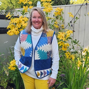 Puede incluir: Un chaleco acolchado con un diseño de patchwork que presenta varios colores, incluyendo azul, turquesa, rosa y amarillo. El chaleco tiene un borde azul marino y se lleva sobre un cuello alto blanco. La persona lleva pantalones amarillos y está de pie frente a flores amarillas.