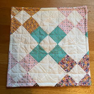 以下が含まれることがあります： 異なる布の切れ端で作られた、ダイヤモンド模様の白いキルト。キルトは、ピンク、イエロー、ターコイズの布でできており、花柄が施されています。