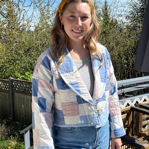 Puede incluir: Una mujer lleva una chaqueta acolchada azul y rosa con cierre de botón. También lleva una diadema amarilla y unos vaqueros azules.