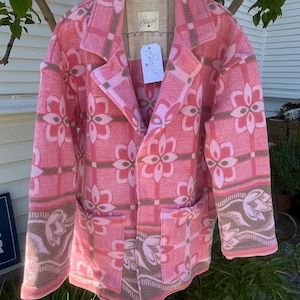 May include: A pink blazer with a floral pattern and two front pockets. The blazer is hanging on a wooden hanger. The tag on the blazer says "Sewirl".