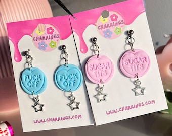 Pode incluir: Brincos com pingentes em forma de coração. Um par é azul claro com a frase "FUCK OFF", o outro rosa com "SUGAR TITS". Cada um tem um berloque de estrela prateada e um pino de bola prateada. O site www.charrings.com é visível.
