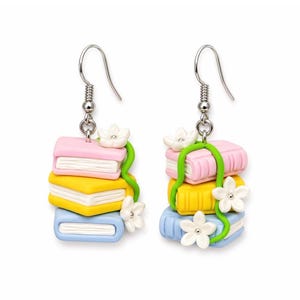 Peut inclure: Boucles d'oreilles fantaisistes représentant des piles de livres roses, jaunes et bleus, ornées de fleurs blanches et d'une vigne verte. Elles sont suspendues à des crochets argentés. Un accessoire charmant et coloré.