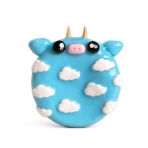 May include: A whimsical, round, light blue cow figurine with white cloud accents. The cow has black eyes, a pink nose, and small golden horns. The figurine is a unique decorative item.