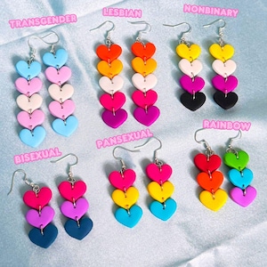Könnte beinhalten: Eine Sammlung farbenfroher herzförmiger Ohrringe. Jedes Paar hat einen anderen Farbverlauf, mit Textbeschriftungen wie "Transgender", "Lesbisch", "Nonbinary", "Bisexuell", "Pansexuell" und "Regenbogen".