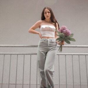 May include: A woman wearing a white crop top, light gray jeans, and red shoes is holding a bouquet of purple flowers. She is standing on a metal railing.