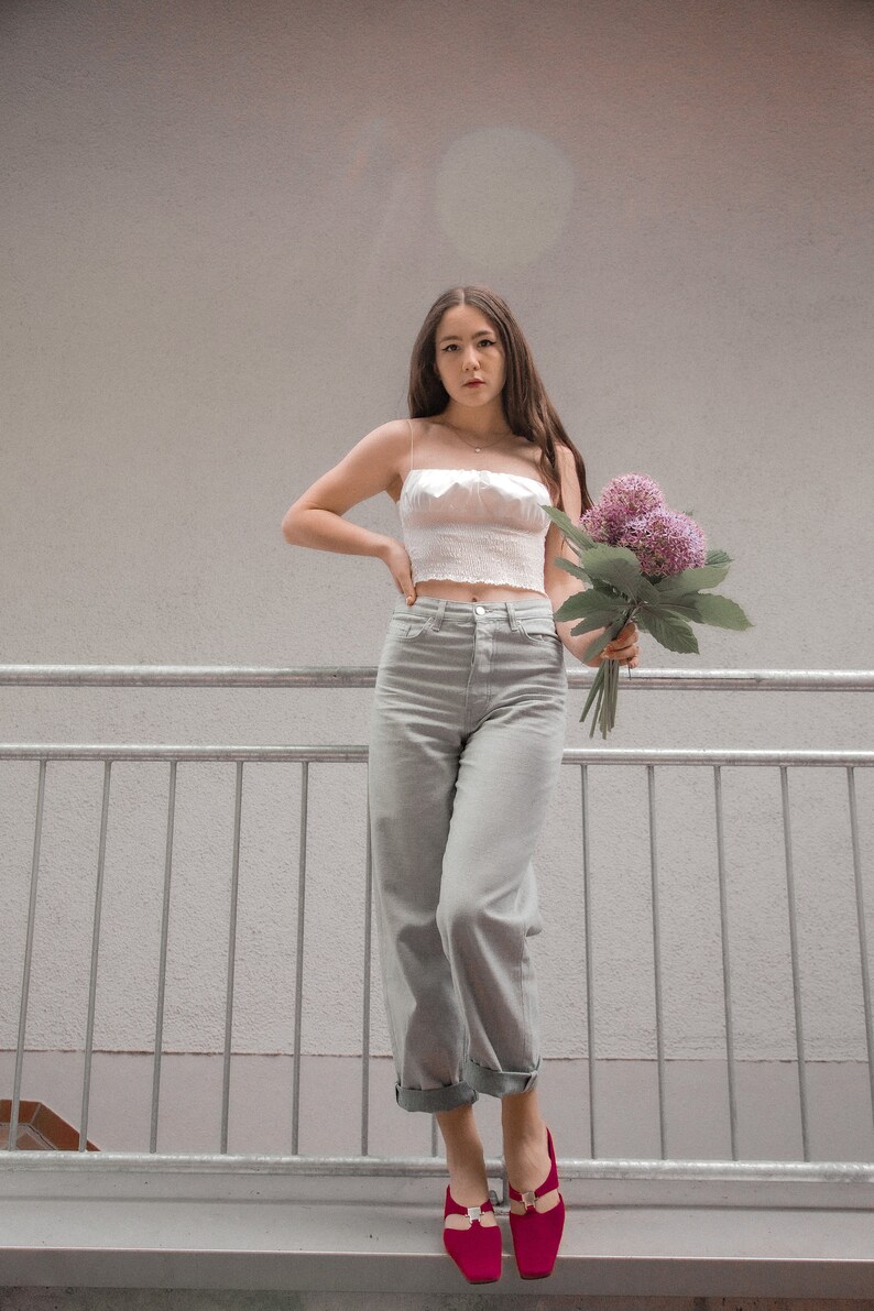 May include: A woman wearing a white crop top, light gray jeans, and red shoes is holding a bouquet of purple flowers. She is standing on a metal railing.