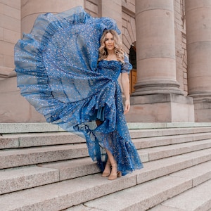 K&ouml;nnte beinhalten: Eine Frau in einem blauen Kleid mit Sternenmuster und einem R&uuml;schen-Saum, die auf einer Steintreppe vor einem Geb&auml;ude mit S&auml;ulen steht.