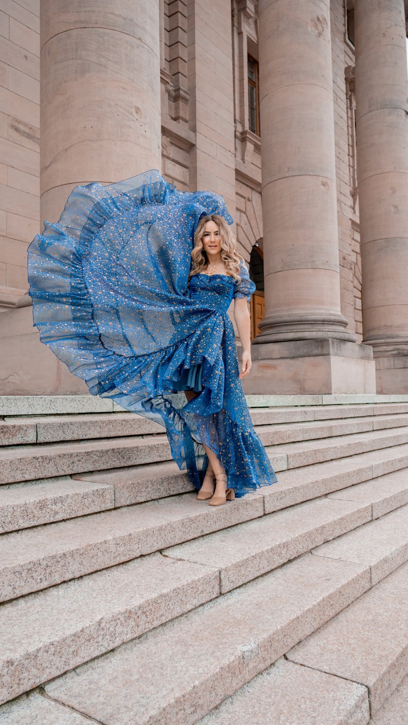 K&ouml;nnte beinhalten: Eine Frau in einem blauen Kleid mit Sternenmuster und einem R&uuml;schen-Saum, die auf einer Steintreppe vor einem Geb&auml;ude mit S&auml;ulen steht.