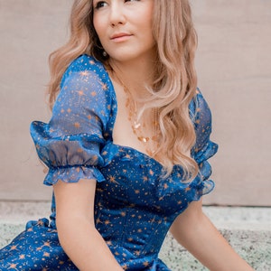 K&ouml;nnte beinhalten: Eine Frau tr&auml;gt ein blaues Kleid mit goldenen Sternen. Das Kleid hat einen herzf&ouml;rmigen Ausschnitt und kurze, bauschige &Auml;rmel. Sie tr&auml;gt eine goldene Halskette mit mehreren Str&auml;ngen.