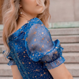 K&ouml;nnte beinhalten: Eine Frau tr&auml;gt ein blaues Kleid mit einem goldenen Sternenmuster. Das Kleid hat einen quadratischen Ausschnitt und Puff&auml;rmel. Die Frau steht vor einer Steintreppe.