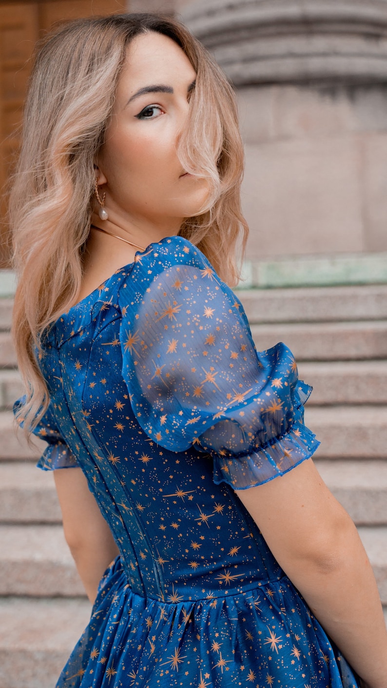 K&ouml;nnte beinhalten: Eine Frau tr&auml;gt ein blaues Kleid mit einem goldenen Sternenmuster. Das Kleid hat einen quadratischen Ausschnitt und Puff&auml;rmel. Die Frau steht vor einer Steintreppe.