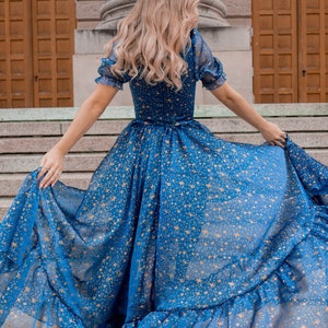 K&ouml;nnte beinhalten: Eine Frau tr&auml;gt ein blaues Maxikleid mit goldenen Sternen. Das Kleid hat einen R&uuml;schen-Saum und ist aus einem durchsichtigen Stoff.