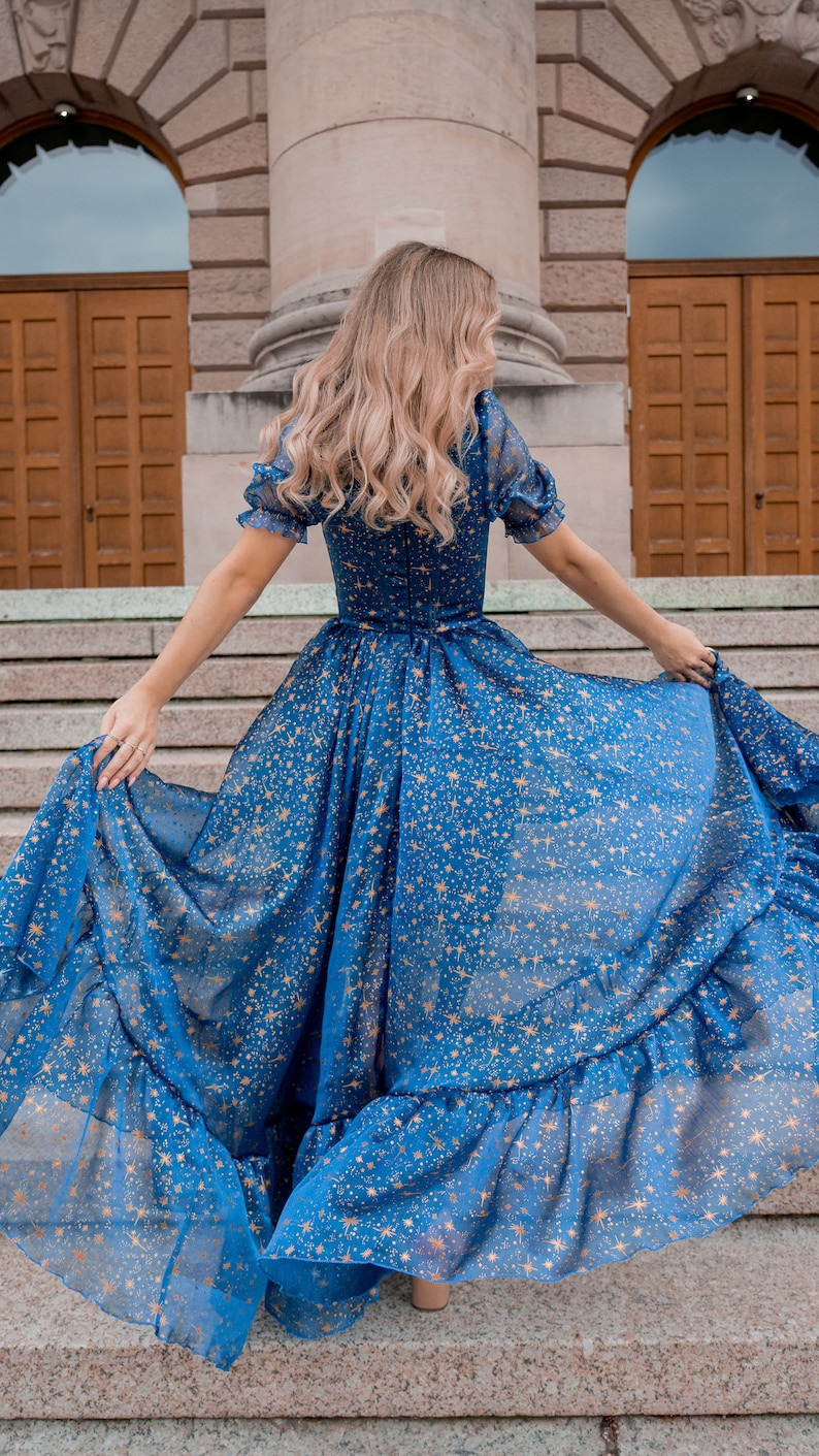 K&ouml;nnte beinhalten: Eine Frau tr&auml;gt ein blaues Maxikleid mit goldenen Sternen. Das Kleid hat einen R&uuml;schen-Saum und ist aus einem durchsichtigen Stoff.