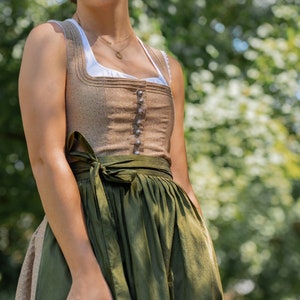 May include: A woman wearing a traditional dirndl dress with a brown bodice and a green skirt. The bodice has a white collar and a row of buttons down the front. The skirt is tied with a brown sash. She is standing in front of a green background.