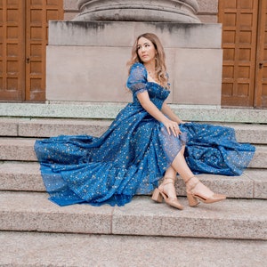 K&ouml;nnte beinhalten: Eine Frau in einem blauen Kleid mit goldenen Sternen bedruckt, sitzt auf einer Steintreppe. Sie tr&auml;gt nude High Heels und hat ihre Haare offen.
