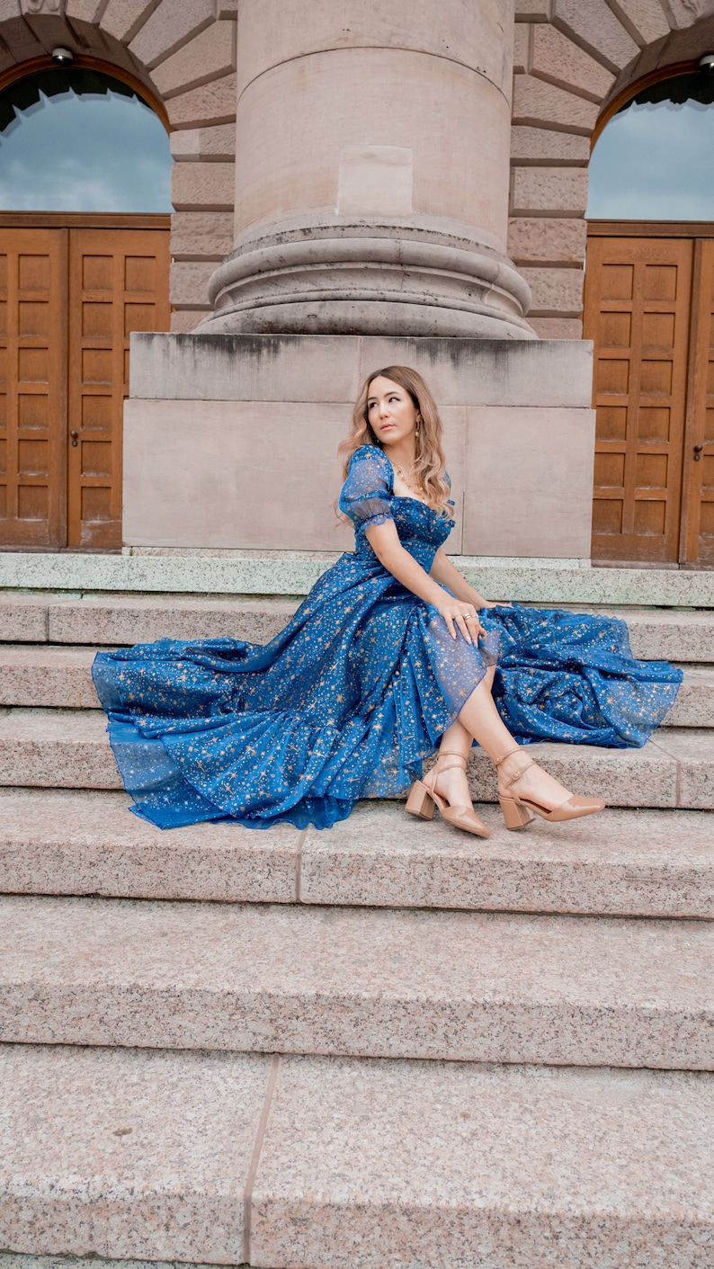 K&ouml;nnte beinhalten: Eine Frau in einem blauen Kleid mit goldenen Sternen bedruckt, sitzt auf einer Steintreppe. Sie tr&auml;gt nude High Heels und hat ihre Haare offen.