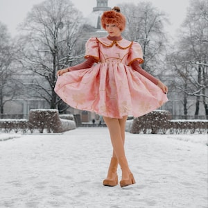 May include: A woman wearing a pink dress with gold embroidery and a brown fur hat stands in a snowy landscape. She is wearing brown heeled boots.