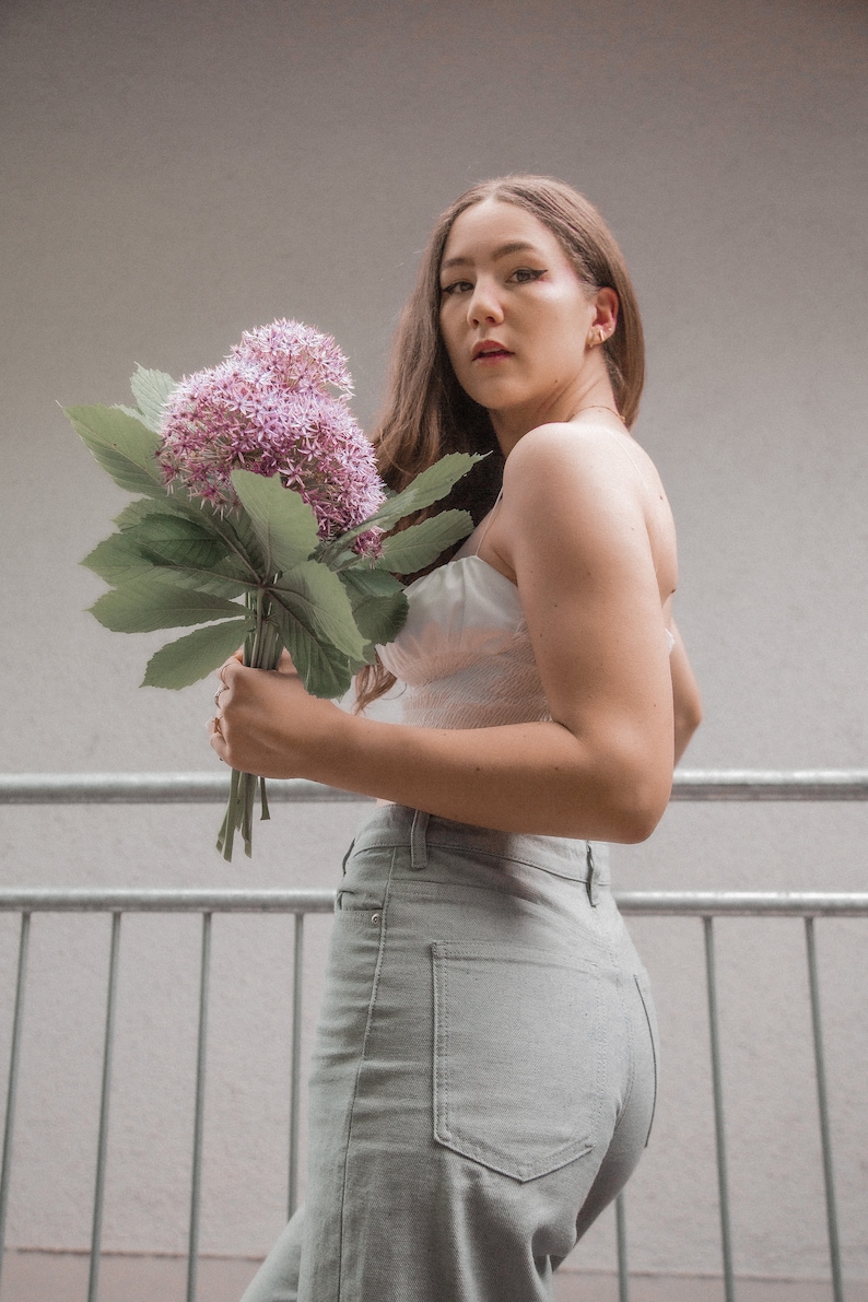 May include: A woman wearing a white crop top and light gray jeans holds a bouquet of purple flowers.