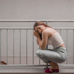 May include: A woman wearing a white crop top and light blue pants sits on a railing with her arms crossed. She is wearing red shoes.