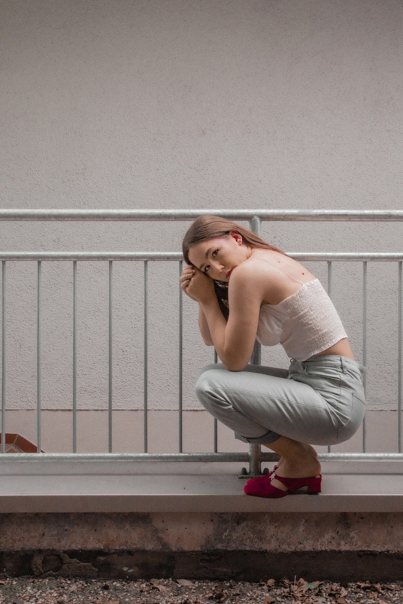 May include: A woman wearing a white crop top and light blue pants sits on a railing with her arms crossed. She is wearing red shoes.