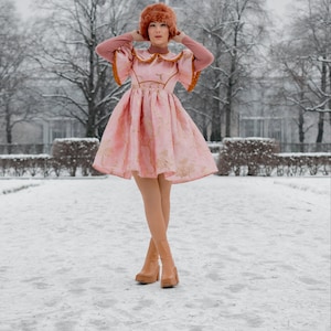 May include: A woman wearing a pink dress with gold embroidery and a brown fur hat stands in a snowy park. She is wearing brown heeled boots.