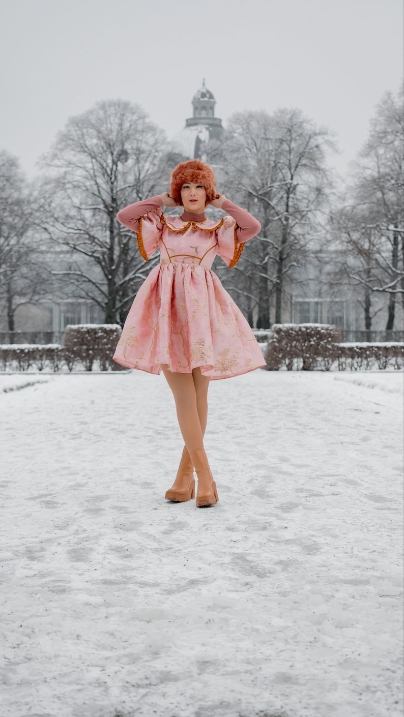 May include: A woman wearing a pink dress with gold embroidery and a brown fur hat stands in a snowy park. She is wearing brown heeled boots.