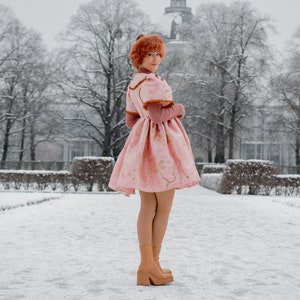 May include: A woman wearing a pink dress with gold floral embroidery and a brown fur hat stands in a snowy park. She is wearing brown heeled boots and looking over her shoulder.