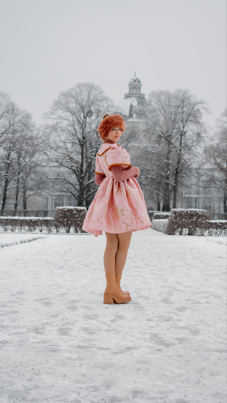 May include: A woman wearing a pink dress with gold floral embroidery and a brown fur hat stands in a snowy park. She is wearing brown heeled boots and looking over her shoulder.