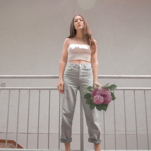 May include: A woman wearing a white crop top and light gray jeans stands in front of a gray railing. She is holding a bouquet of purple flowers. She is wearing red shoes with a white buckle.