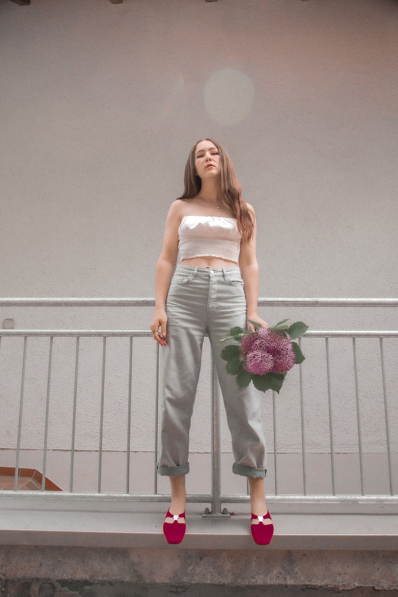 May include: A woman wearing a white crop top and light gray jeans stands in front of a gray railing. She is holding a bouquet of purple flowers. She is wearing red shoes with a white buckle.