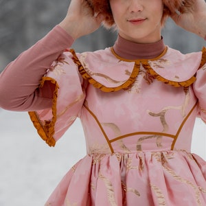 May include: A woman wearing a pink dress with gold embroidery and a brown fur hat. The dress has a large collar with a ruffle and a fitted bodice. The skirt is full and flowing.