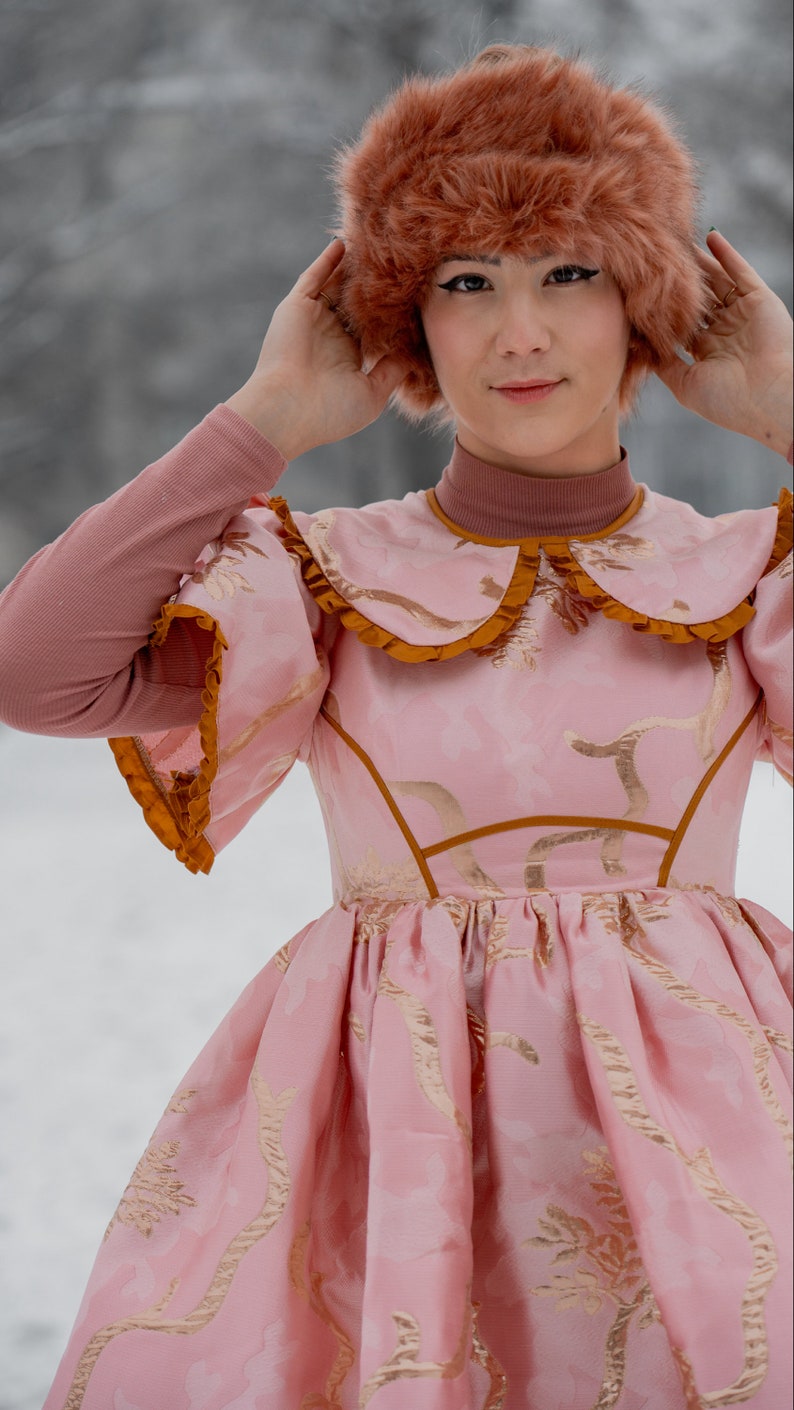 May include: A woman wearing a pink dress with gold embroidery and a brown fur hat. The dress has a large collar with a ruffle and a fitted bodice. The skirt is full and flowing.