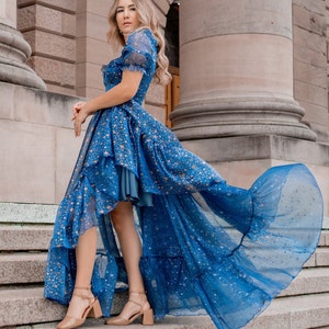 K&ouml;nnte beinhalten: Eine Frau tr&auml;gt ein blaues Kleid mit goldenen Sternenmuster und einem hohen, niedrigen Saum. Das Kleid hat einen R&uuml;schenrock und ist aus einem durchsichtigen Stoff gefertigt. Die Frau steht auf einer Treppe.