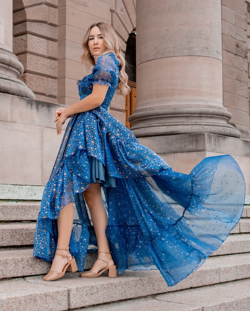 K&ouml;nnte beinhalten: Eine Frau tr&auml;gt ein blaues Kleid mit goldenen Sternenmuster und einem hohen, niedrigen Saum. Das Kleid hat einen R&uuml;schenrock und ist aus einem durchsichtigen Stoff gefertigt. Die Frau steht auf einer Treppe.