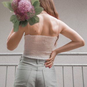 May include: A person wearing a white crop top and light gray wide-leg pants. They are holding a bouquet of pink flowers behind their back.